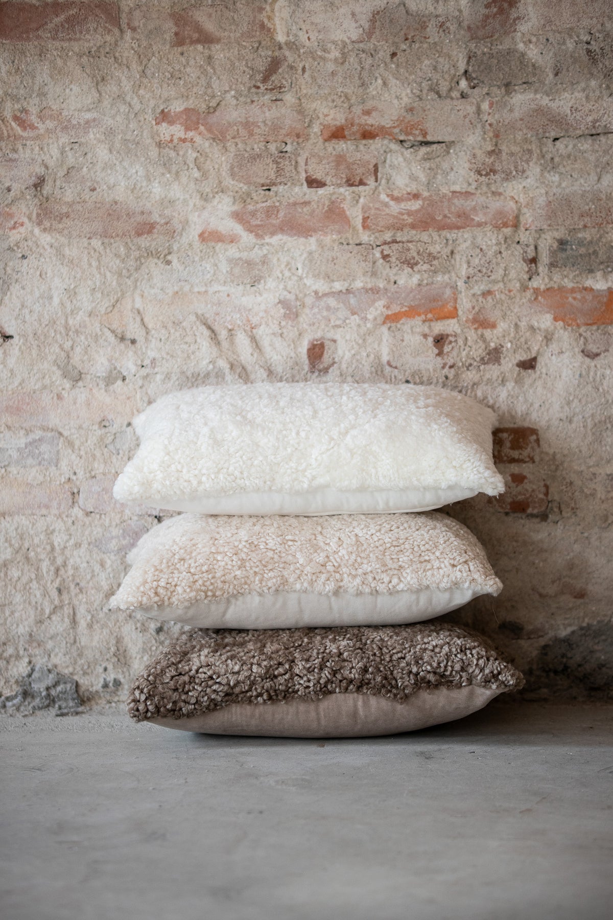 Pile de coussins en peau de mouton bouclée blanc, clair de lune et sahara devant un mur de briques