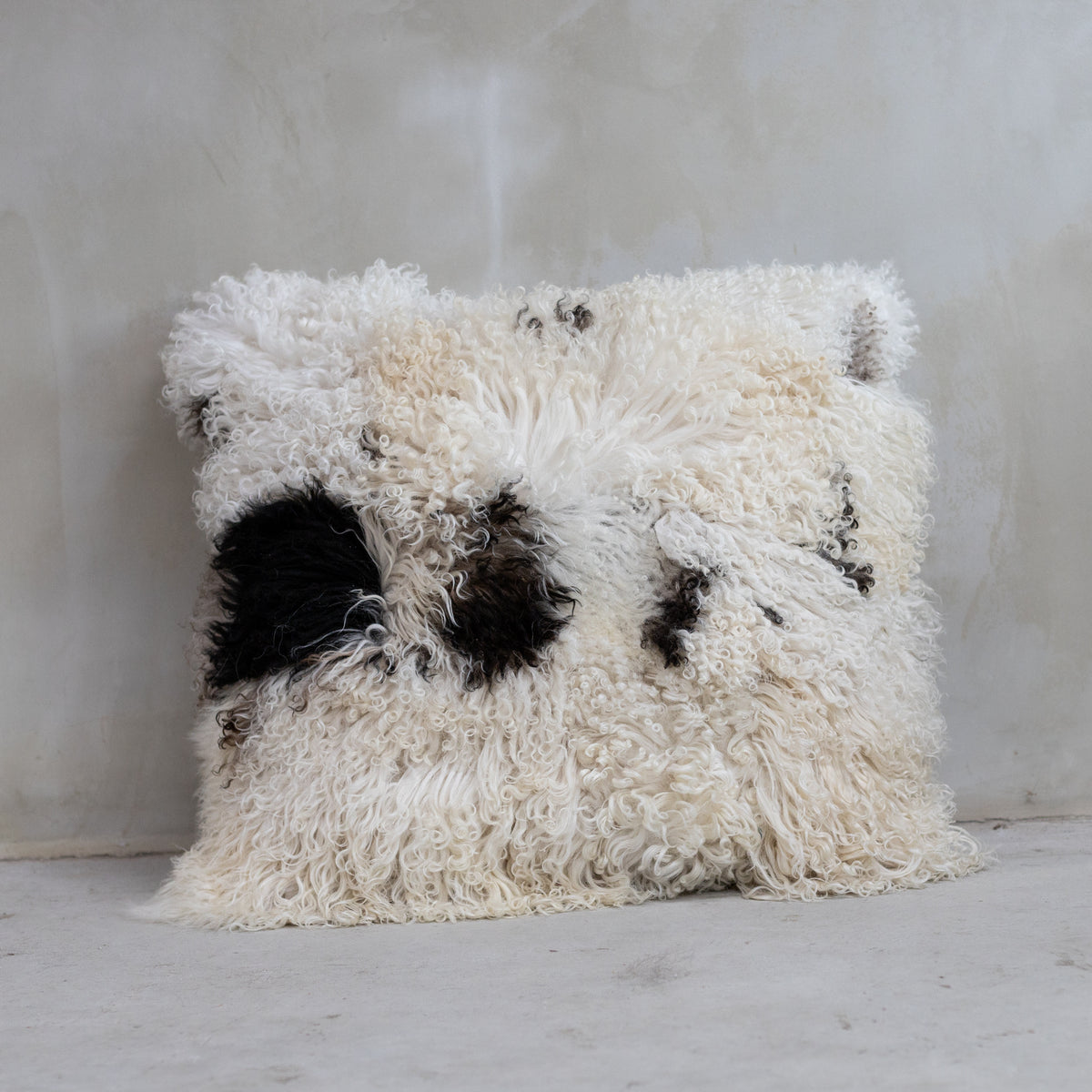 Coussin XXL 80x80 cm en peau de mouton bouclée crème, brune et noire posé contre un mur